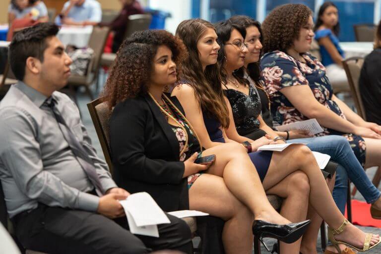 Diverse students of color in formal wear sit in a row listening to speaker